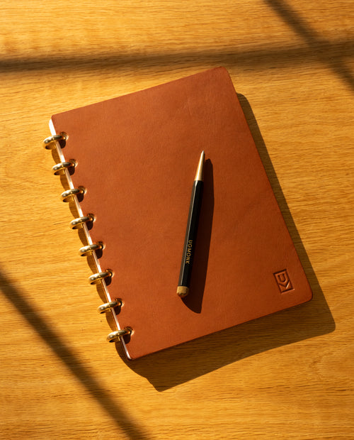 A Discbound Heirloom Journal + 3 Refills (Tan Leather - Standard 6″ x 8.5″) with elegant gold rings and a black pen placed on top rests on a wooden surface, sunlight casting gentle shadows across the scene.