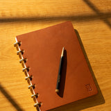 The Discbound Heirloom Journal in tan leather (Standard 6" x 8.5") rests on a wooden surface with diagonal shadows, gold rings glinting and a black and gold pen placed neatly on top.