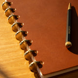 A Discbound Heirloom Journal in tan leather (standard 6" x 8.5”) with dot grid pages and a pen rests elegantly on the table.