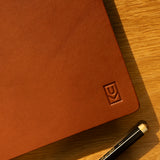 A Discbound Heirloom Journal (Tan Leather - Standard 6" x 8.5") with an embossed logo sits on a wooden surface next to a black pen with gold accents, bathed in sunlight and warm shadows.