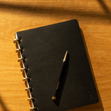 A Discbound Heirloom Journal (Black Leather - Standard 6" x 8.5") and a matching black pen rest on a wooden surface, bathed in warm sunlight and soft shadows.