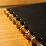 A close-up of the Discbound Heirloom Journal (Black Leather - Standard 6" x 8.5") with gold disc binding rings, resting on a wooden surface in warm sunlight, highlighting the edge and elegant, refillable rings.