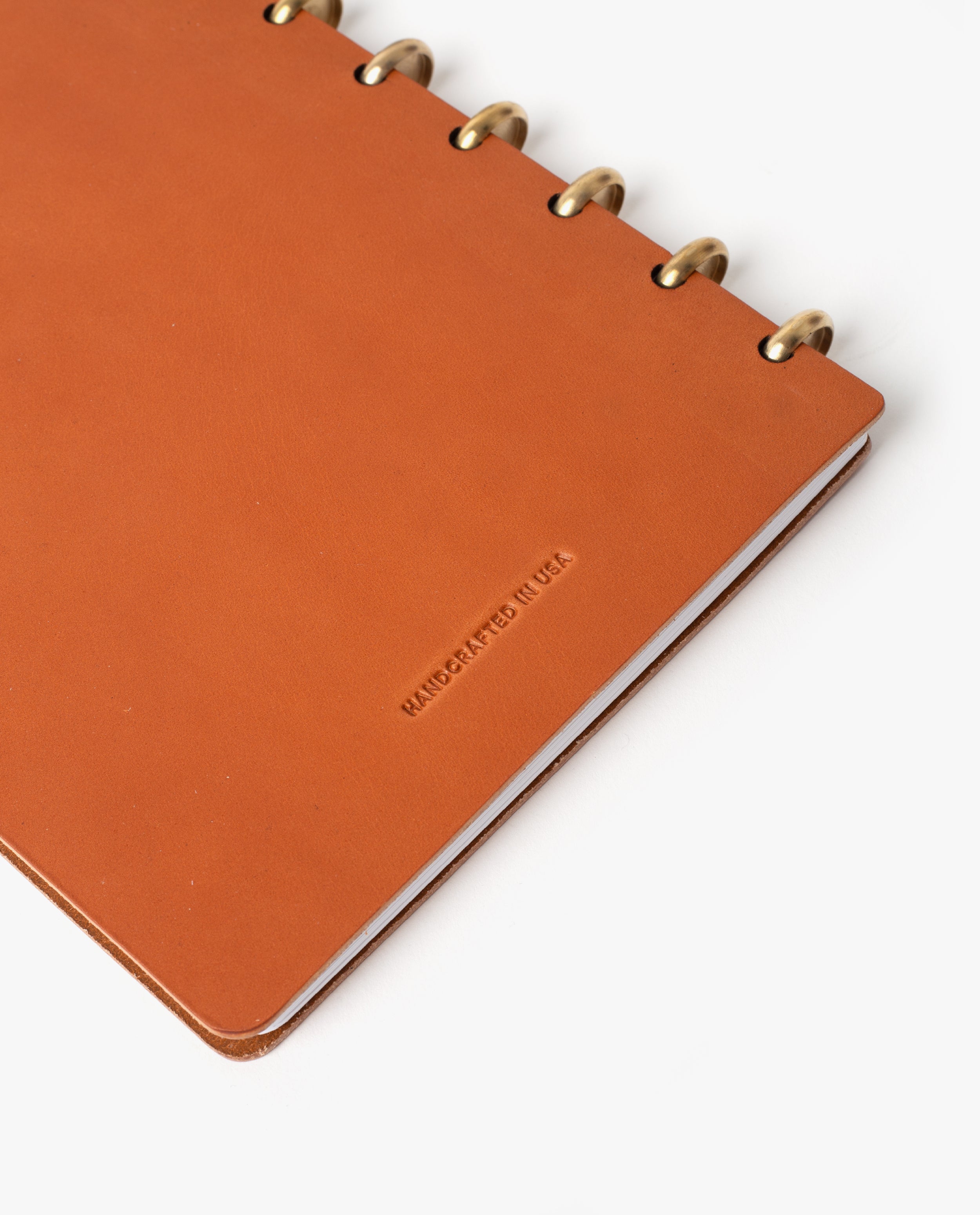 A tan, full-grain leather Discbound Heirloom Journal Bundle (Standard 6" x 8.5" + 3 Refills) with brass rings rests partially open on a white background. The cover is stamped with HANDCRAFTED IN USA in small letters, emphasizing the refined quality of this elegant refillable discbound notebook.