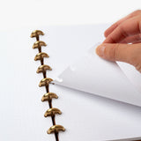 A hand lifts a dotted page from the Discbound Heirloom Journal (Tan Leather - Standard 6" x 8.5") with gold disc binding, featuring removable and repositionable dot grid paper against a white background.