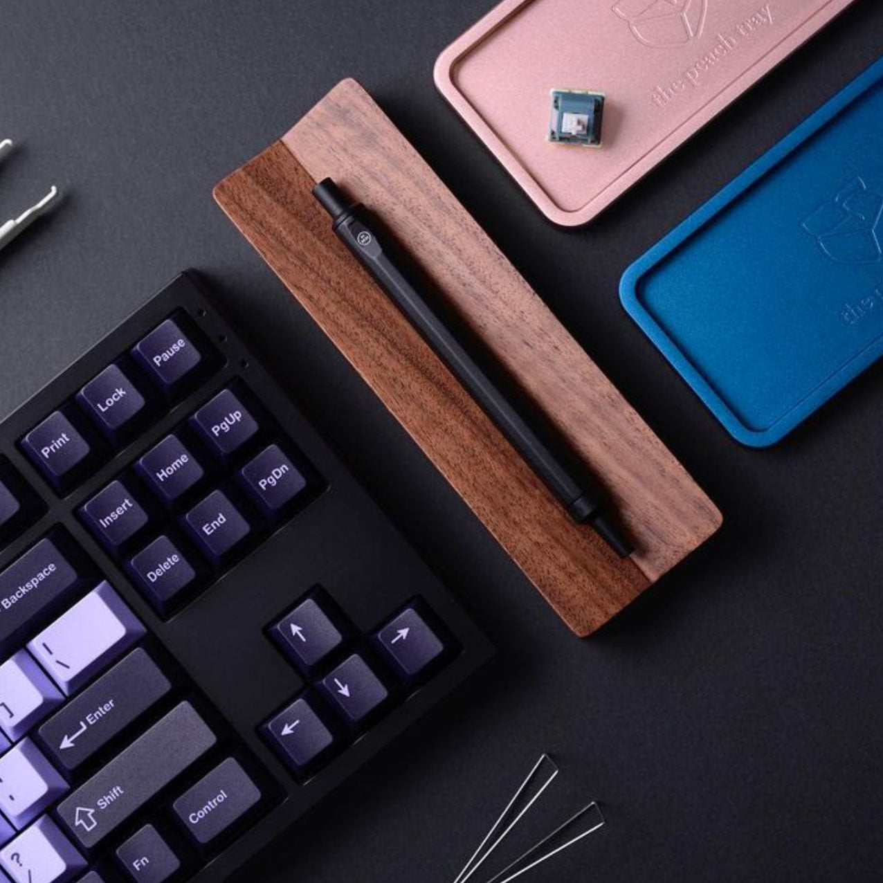 A mechanical keyboard with black and white keys sits next to a wooden pen holder with a black pen, blue and pink trays, a switch, and keyboard tools on a dark surface.