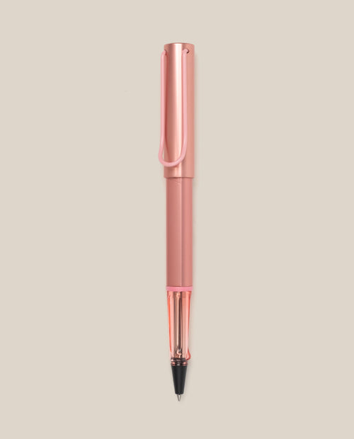 The LAMY AL-star Rollerball (Flamingo) features a pink anodized aluminum body, clear grip, and black ink tip, shown vertically on a light beige background for smooth writing.