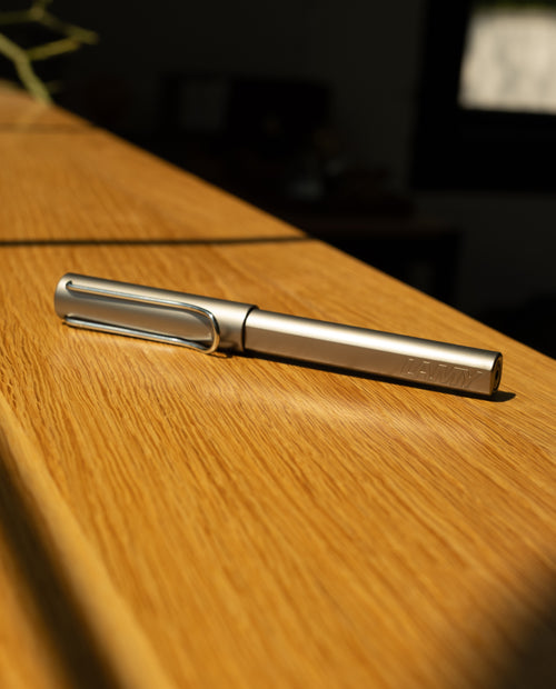A graphite LAMY AL-star Rollerball lies diagonally on a sunlit wooden surface, with natural light casting shadows and highlighting its metallic finish, offering smooth writing performance.