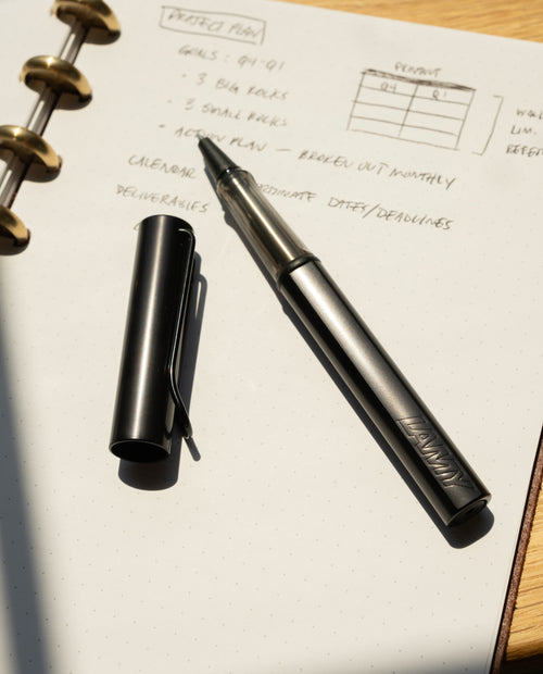 A LAMY AL-star Rollerball (Black) with its cap off rests on an open dotted notebook showing handwritten project notes. Sunlight casts shadows across the page, highlighting the pen’s smooth writing experience.