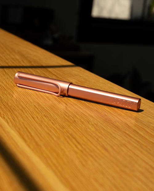 The LAMY AL-star Rollerball (Flamingo), featuring a metallic pink anodized aluminum body, lies on a wooden surface in sunlight, casting gentle shadows and highlighting its modern design.