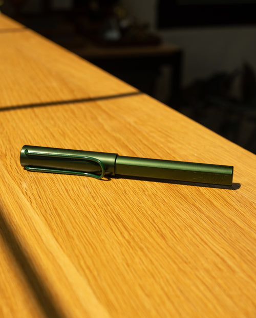 A LAMY AL-star Rollerball (Pine) rests on a sunlit wooden surface, casting a rightward shadow—a refillable rollerball pen recognized for its smooth writing experience.