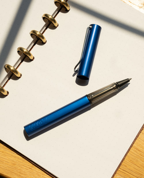 A LAMY AL-star Rollerball (Oceanblue) with its cap off rests on an open dotted notebook with gold rings, set on a wooden surface in bright sunlight, highlighting its smooth writing and refillable rollerball design.