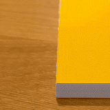 A hand taps the top corner of a yellow-covered Layflat Notebook Bundle (Set of 3) resting on a wooden table, making the notebook’s corner bounce slightly.