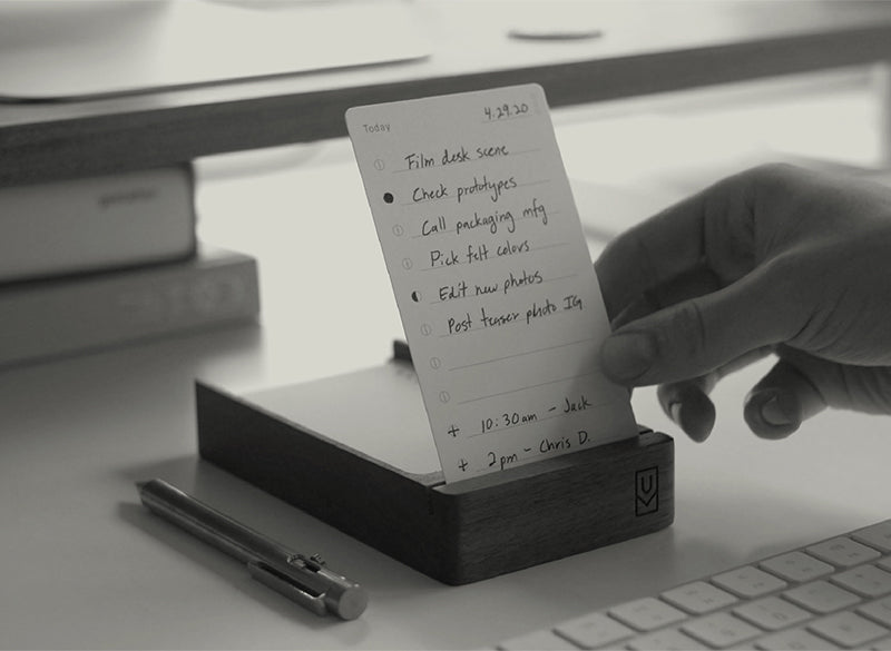 A hand holds a notecard with a handwritten to-do list, placed on a wooden holder beside a pen, keyboard, and closed laptop on a white desk. The list includes tasks and two appointments.