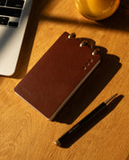 A Leather Discbound Analog (Brown) notepad, a black pen labeled UCONN, and part of a laptop rest on a wooden desk in warm sunlight.