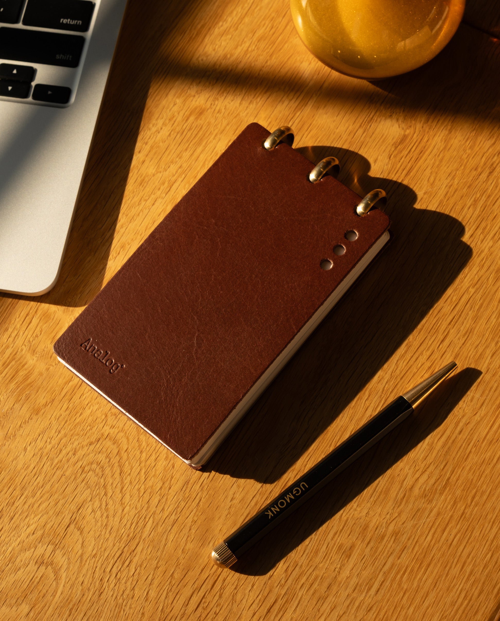 A Leather Discbound Analog (Brown) notepad, a black pen labeled UCONN, and part of a laptop rest on a wooden desk in warm sunlight.