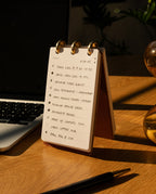 A Leather Discbound Analog + 3 Refills (Tan) rests on the desk beside a laptop and an hourglass, as sunlight casts shadows across the wooden surface. Inside, to-do lists with tasks like team call, script rewrite, and emailing merge classic elegance with everyday productivity.