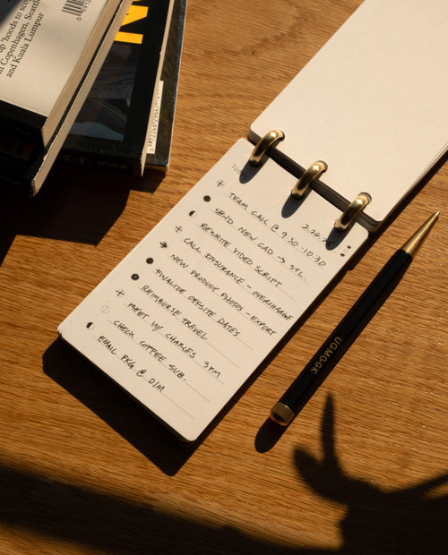 A notepad with a handwritten to-do list and a black and gold pen rest on a wooden desk beside stacked books in warm sunlight, casting shadows across the scene. The presence of the Leather Discbound Analog (Tan), featuring full-grain leather covers, adds a touch of sophistication and timeless organization to the workspace.