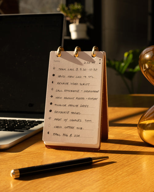 A Leather Discbound Analog (Brown) notepad with a handwritten to-do list sits on a wooden desk next to a black pen, bathed in warm sunlight. The list, organized on Analog Task Cards, includes tasks like team call, rewrite video script, and email FAQ.