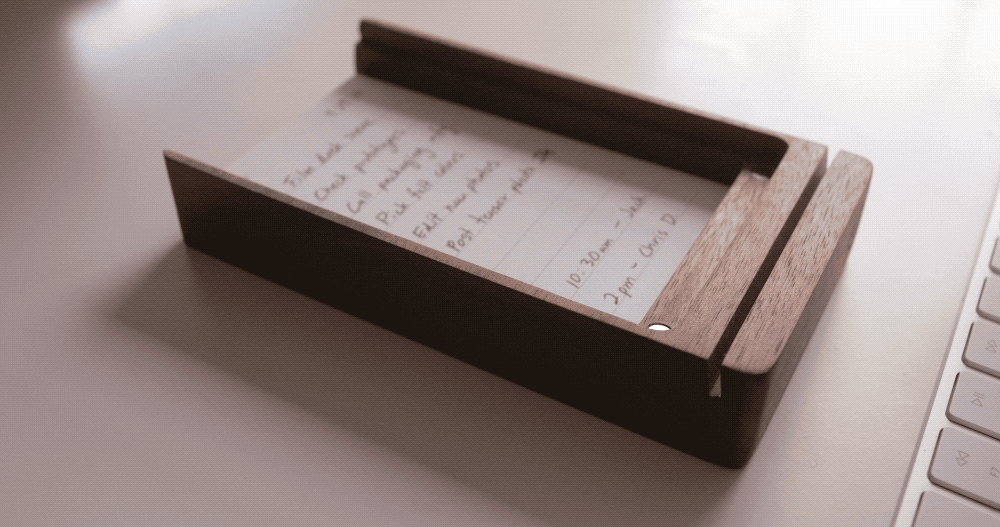 A handwritten to-do list on notepaper sits in a wooden holder beside a white computer keyboard on a desk.