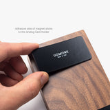 A hand attaches the Magnet Kit (For Analog Card Holder)—a black strip labeled UGMONK Made in USA—to a wooden Analog Card Holder with strong adhesive magnets. Text above: Adhesive side of magnet sticks to the Analog Card Holder.