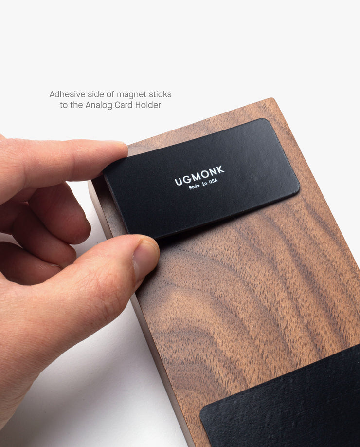 A hand attaches the Magnet Kit (For Analog Card Holder)—a black strip labeled UGMONK Made in USA—to a wooden Analog Card Holder with strong adhesive magnets. Text above: Adhesive side of magnet sticks to the Analog Card Holder.