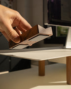 A hand places or removes a small wooden object from the Warehouse Item - Magnet Kit (Pen Tray and Analog Weekly) on a light-colored desk with wooden legs. A computer screen and the sleek Gather Monitor Stand are visible in the background.