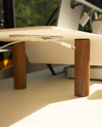 A close-up of a desk with wooden legs features a gray felt pad and a white charging cable, neatly managed with Warehouse Item - Magnetic Cable Keepers (Set of 2 - Light Gray). Sunlight streams in, casting warm shadows across the scene, while computer equipment and greenery are visible in the background.