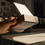A hand holds a Magnet Kit for Analog Card Holder with a notecard inside, angled toward sunlight on a white desk. The kit features strong adhesive magnets for stability and a small logo engraved on the side.