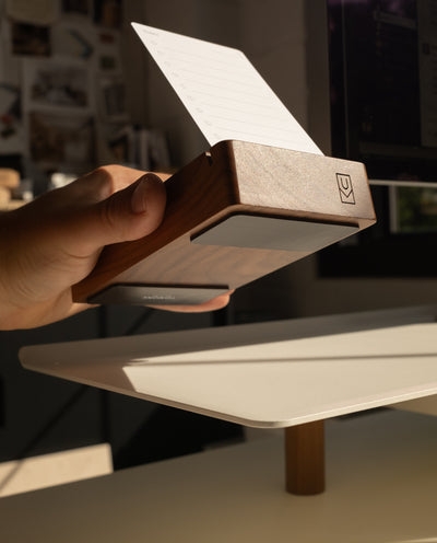 A hand holds a Magnet Kit for Analog Card Holder with a notecard inside, angled toward sunlight on a white desk. The kit features strong adhesive magnets for stability and a small logo engraved on the side.