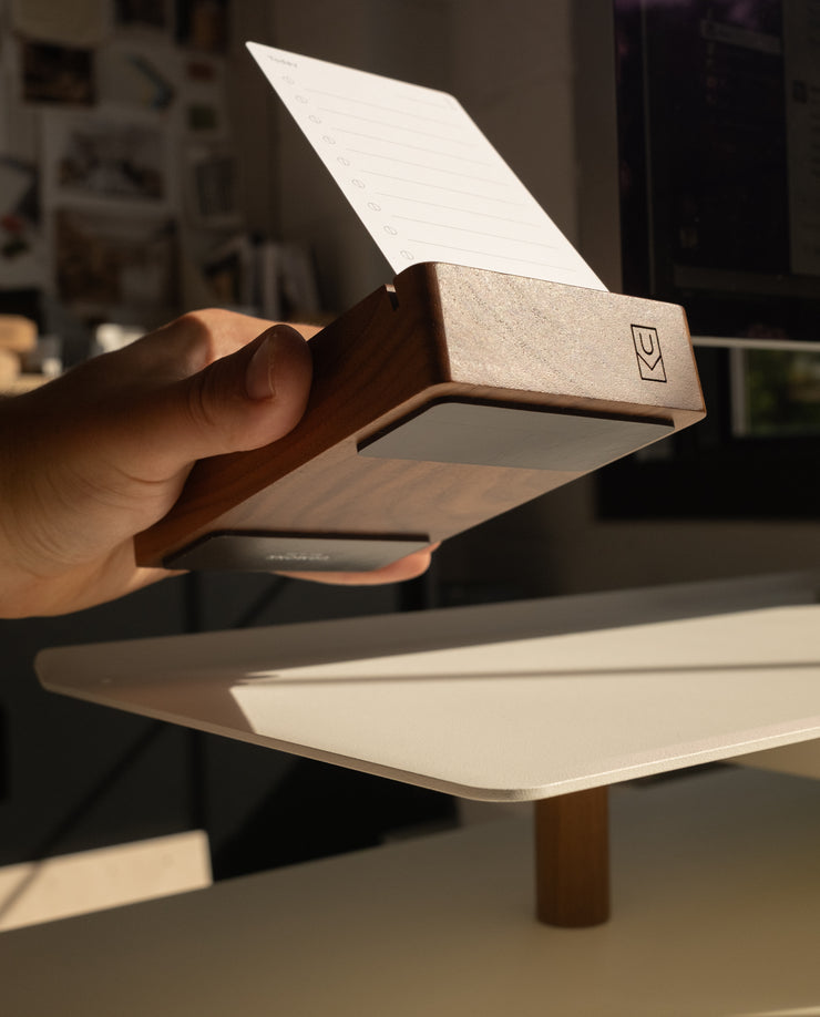 A hand holds a Magnet Kit for Analog Card Holder with a notecard inside, angled toward sunlight on a white desk. The kit features strong adhesive magnets for stability and a small logo engraved on the side.