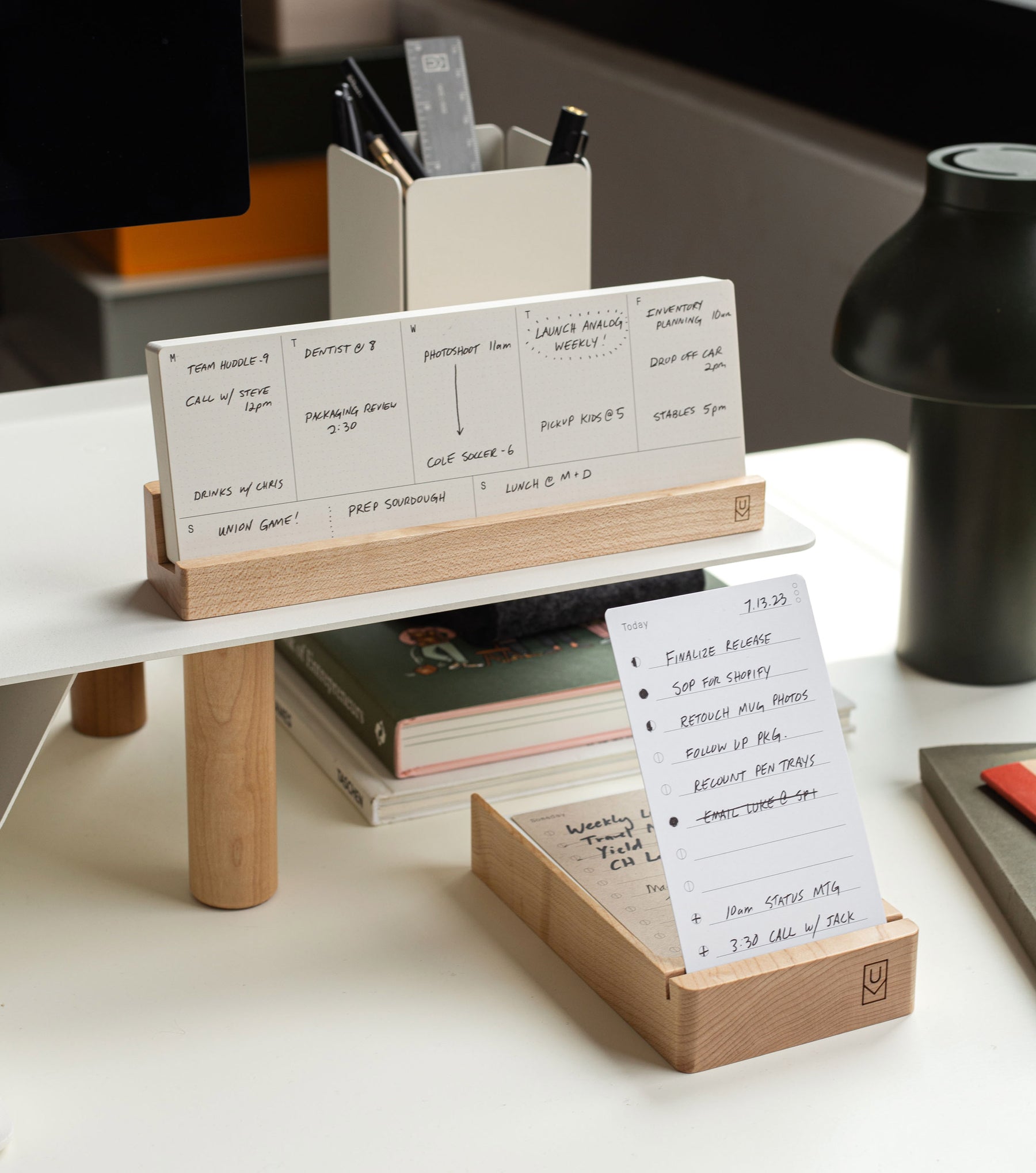 A wooden desk organizer holds notecards and pens beneath a monitor stand. One notecard displays a handwritten daily schedule. Another wooden holder contains more notecards and a small stack of paper on a modern, tidy desk.