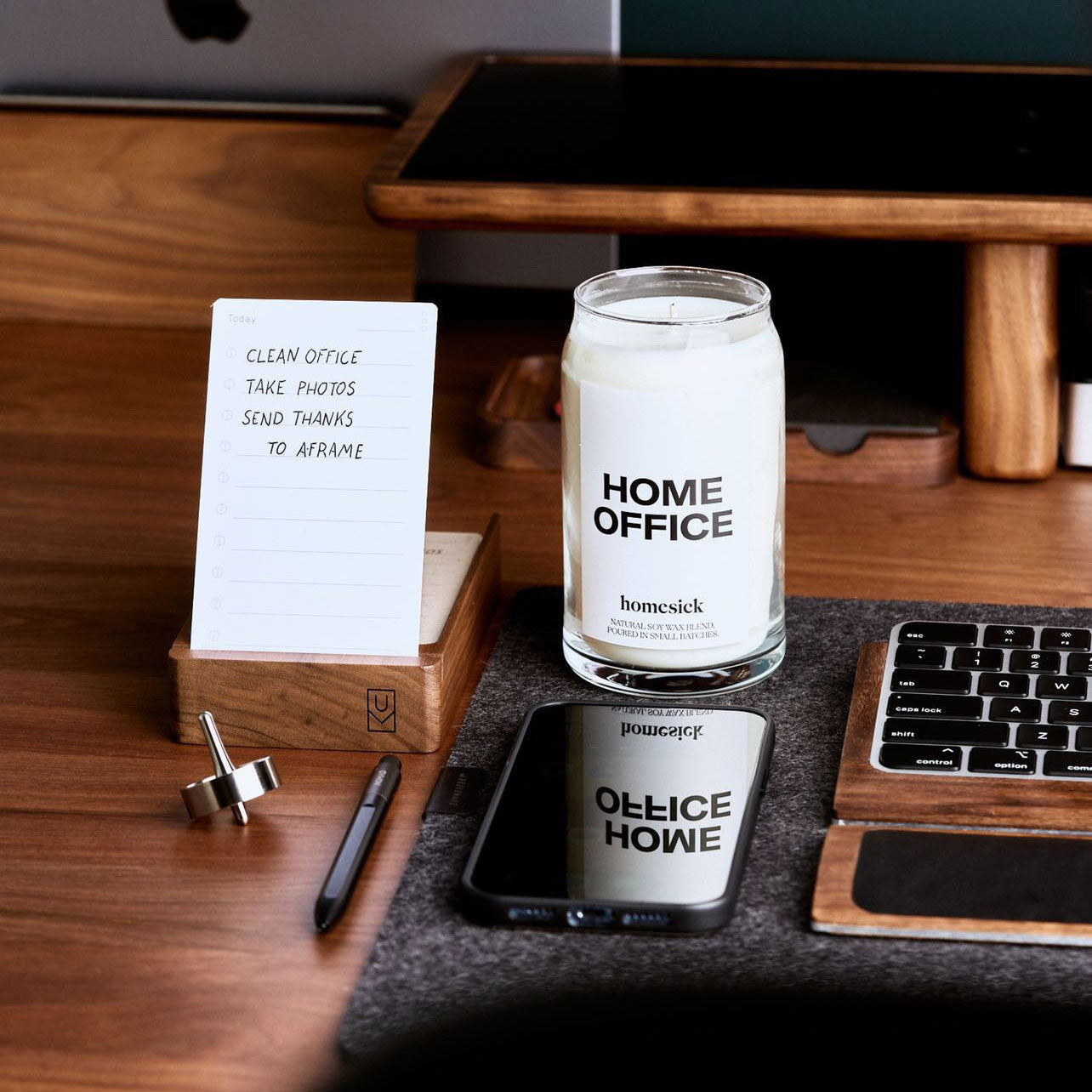 A desk with a notepad listing office tasks, a Home Office candle, a pen, a smartphone, and part of a keyboard and monitor, all arranged neatly on a wooden surface.