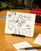 An Analog Starter Kit (Black) card—featuring black ink sketches of stackable containers—rests in its aluminum card stand on a desk beside a red typewriter, a plant, and handwritten notes, evoking the charm of a paper card system.