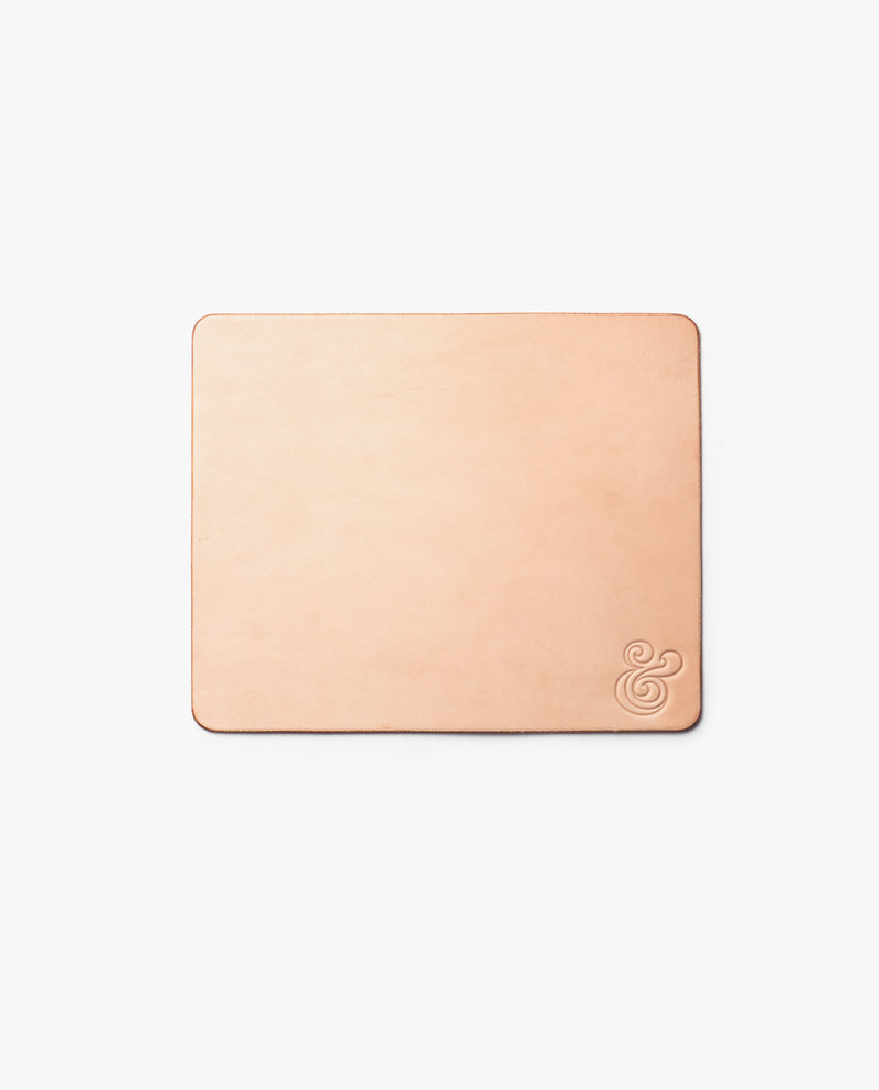 A square, tan leather coaster with rounded edges and an embossed ampersand symbol in the lower right corner, on a plain white background.