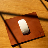 A white wireless mouse sits on the Ugmonk Leather Mousepad (Tan) atop a wooden surface, diagonal shadows falling across the full-grain leather.