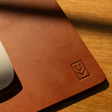 A close-up of the Ugmonk Leather Mousepad (Tan), crafted from brown leather with a geometric logo embossed in the corner, rests on wood. A white computer mouse is partly visible as sunlight casts gentle shadows across the scene.
