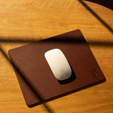 A white Apple Magic Mouse rests on the Ugmonk Leather Mousepad (Brown), handcrafted and set on a wooden desk where diagonal shadows fall across the surface.