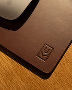 A close-up of the Ugmonk Leather Mousepad (Brown) with an embossed logo in the corner, placed on a wooden surface. The edge of a white computer mouse appears at the top left.