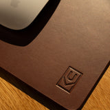 A close-up of the Ugmonk Leather Mousepad (Brown) with an embossed logo in the corner, placed on a wooden surface. The edge of a white computer mouse appears at the top left.