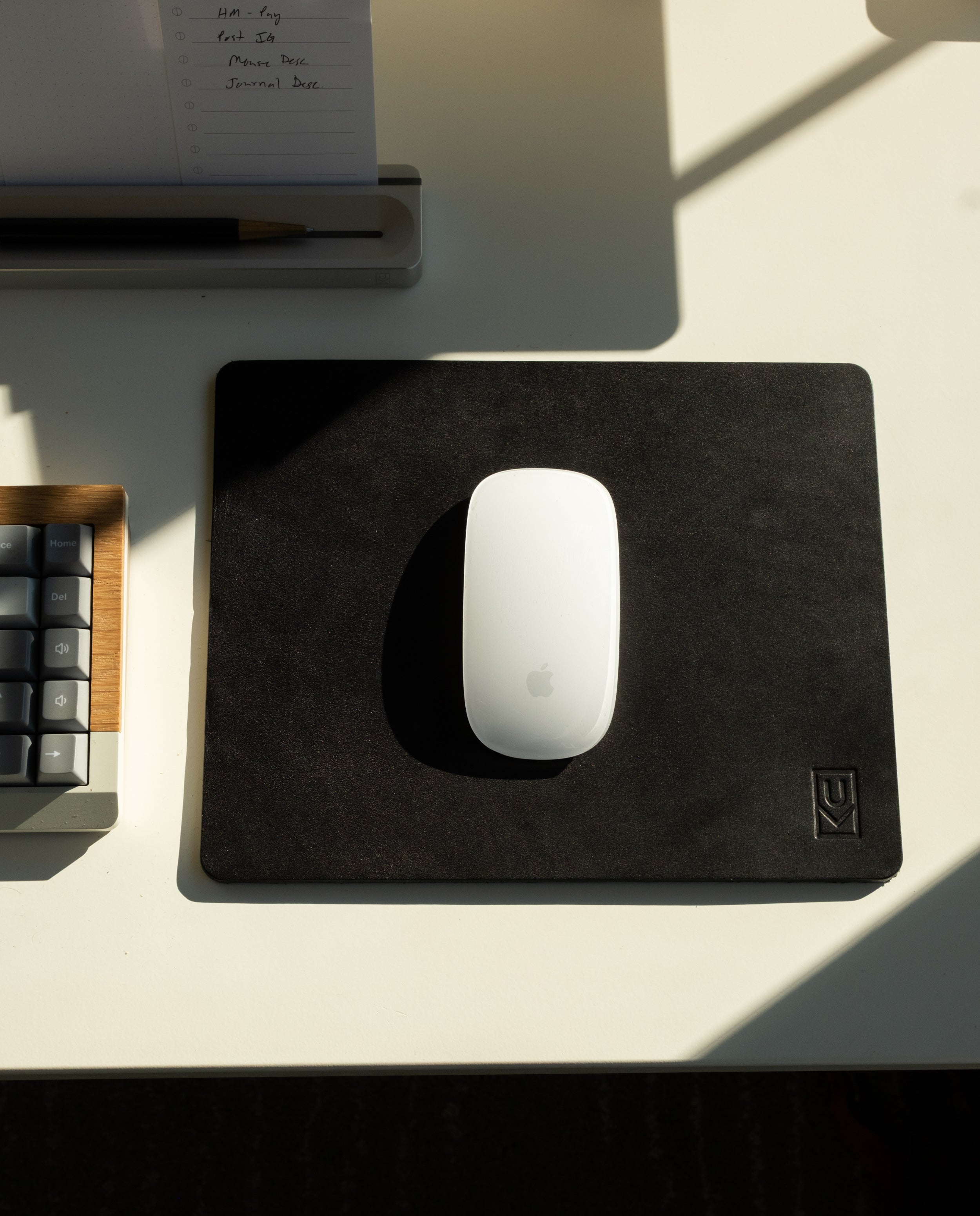 A white Apple Magic Mouse rests on an Ugmonk Leather Mousepad (Black) atop a white desk, next to a partially visible keyboard and a notepad, with sunlight casting shadows across the surface.