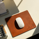 A white wireless mouse sits on the Ugmonk Leather Mousepad (Tan) on a white desk, next to a gray keyboard and notepad. Sunlight casts gentle shadows across the workspace.
