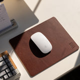A white Apple Magic Mouse sits on a brown Ugmonk Leather Mousepad, next to a gray keyboard and notepad on a sunlit white desk.