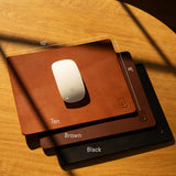 The Ugmonk Leather Mousepad (Tan), crafted from full-grain leather and sized 10" x 8", is shown on a wooden surface beside a white Apple Magic Mouse, catching sunlight and casting elegant shadows.