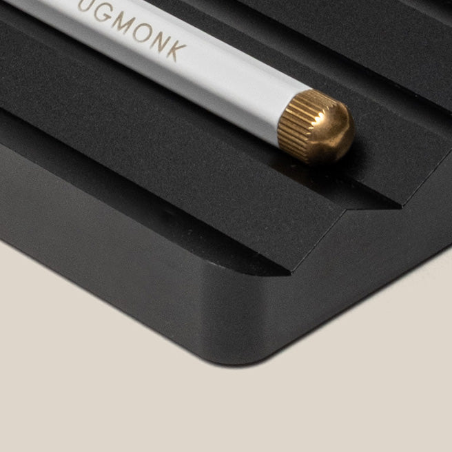Close-up of a white pencil with a gold eraser resting in a grooved, black desk organizer on a beige surface. The pencil has text that reads UGMONK near the eraser end.