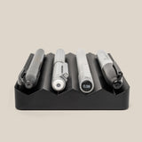 The Complete Analog Collection (Black Aluminum), featuring four uniquely designed pens, is displayed horizontally in individual grooves of a black desktop organizer against a plain beige background.