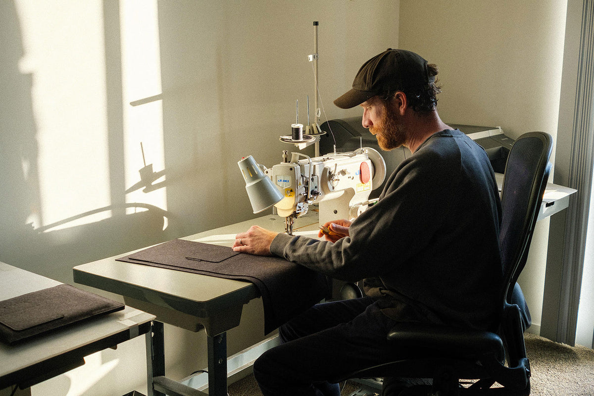 A man wearing a cap sits at a sewing machine, stitching dark fabric in a sunlit room with beige walls. Sunlight casts shadows through a window, creating a warm atmosphere.