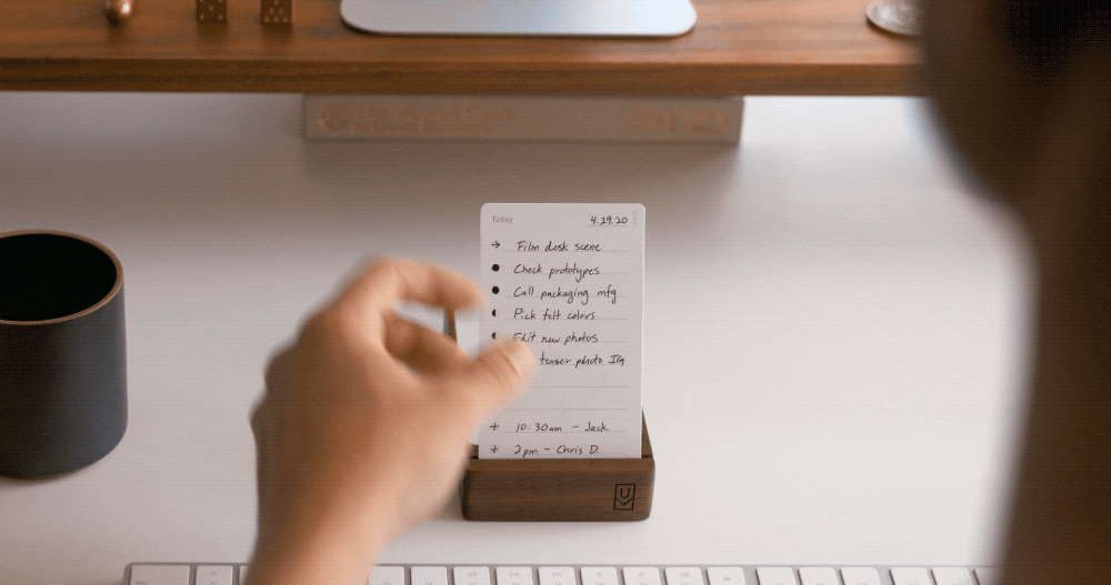 A hand flips a small paper card on a wooden desk stand, revealing a handwritten to-do list. A keyboard, a cup, and a monitor stand are visible in the background on a tidy workspace.