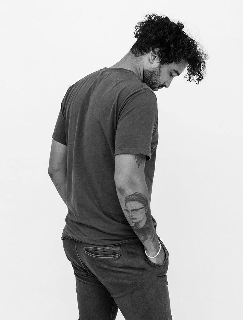 A man with curly hair wearing a plain t-shirt and jeans stands with his back turned, looking down. He has one hand in his pocket and a detailed portrait tattoo on his forearm. The background is plain and white.