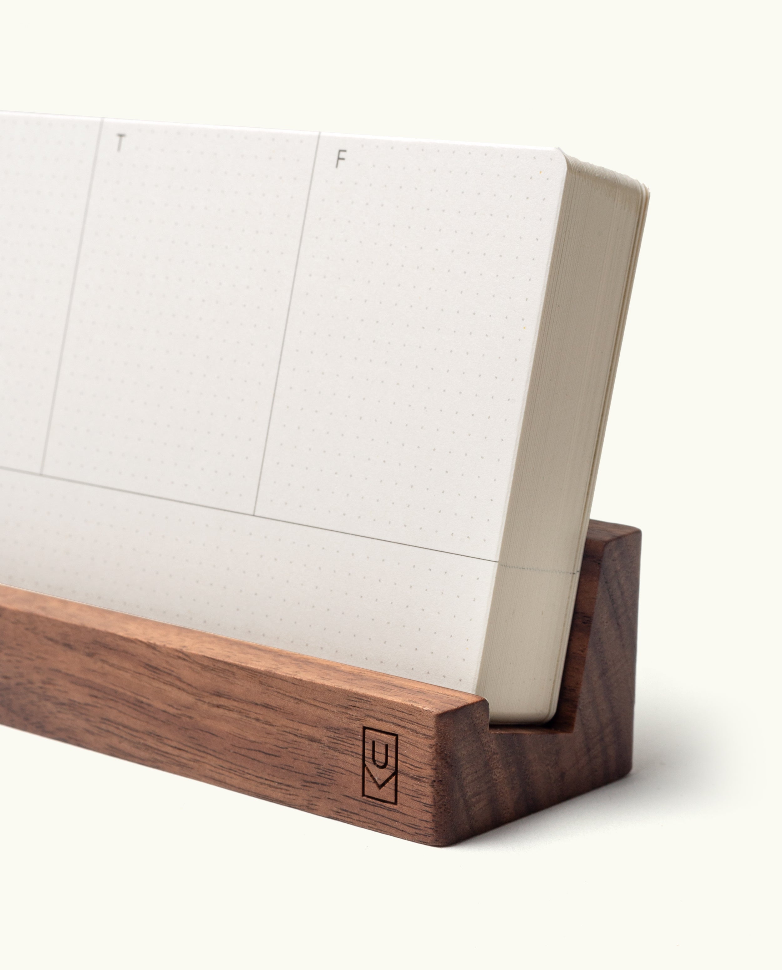 A close-up of the Analog Weekly Planning Kit (Walnut) highlights its dotted weekly planner cards, neatly displayed upright in a minimalist walnut holder with a small engraved logo on the front. Only a portion of the planner is visible.
