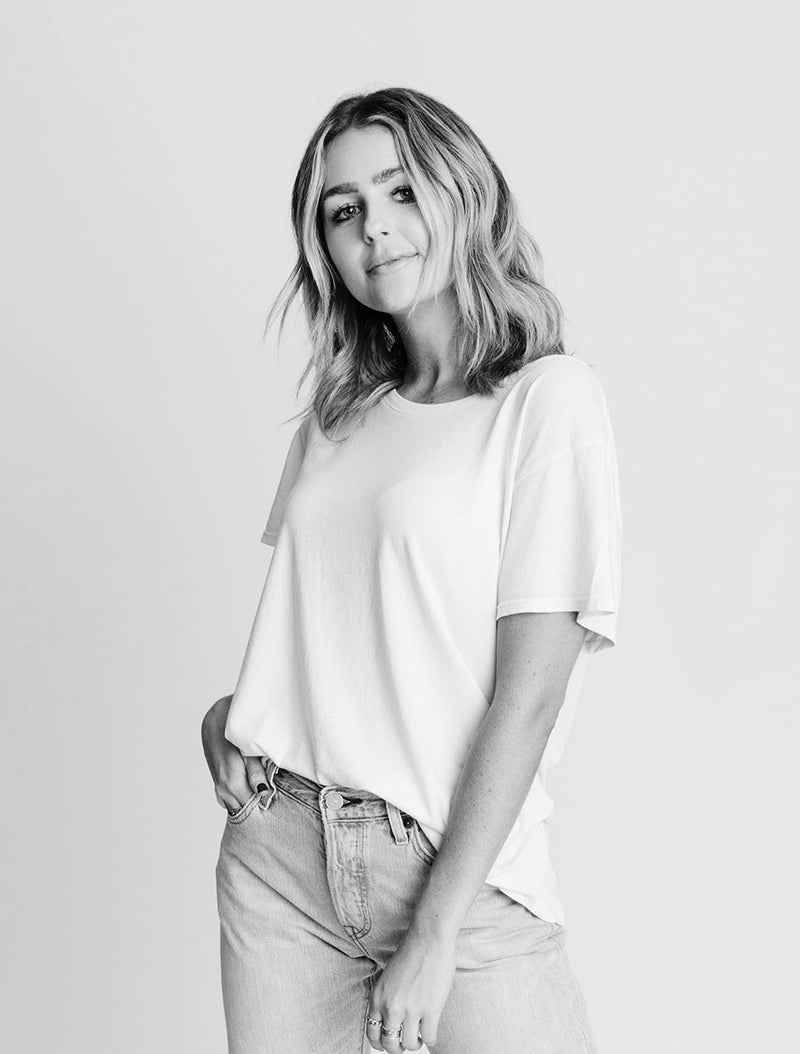 A woman with wavy, shoulder-length hair stands facing the camera, wearing a loose white t-shirt and light jeans. She has one hand in her pocket and is smiling softly against a plain background.